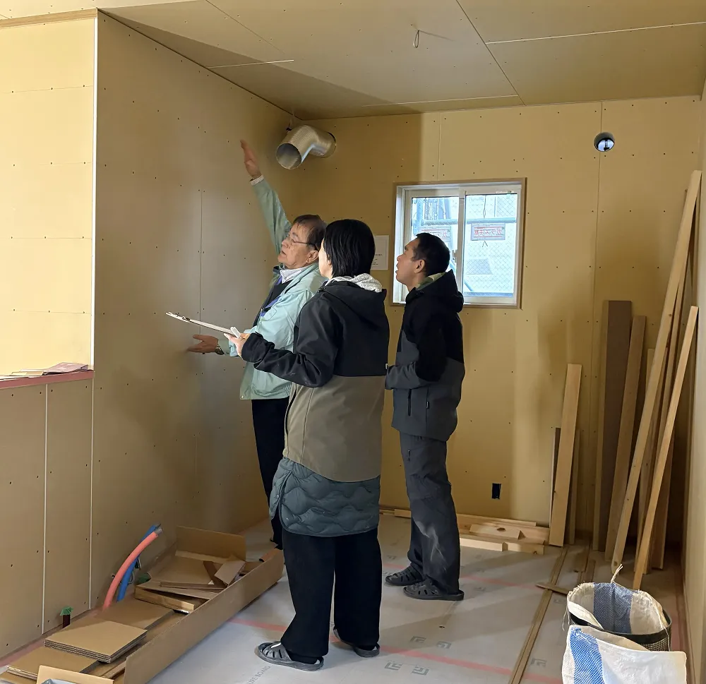 工事現場で天井・壁を指し示しながら打合せする3人の男性（メジャーやタブレットを持った施工・検査の様子）