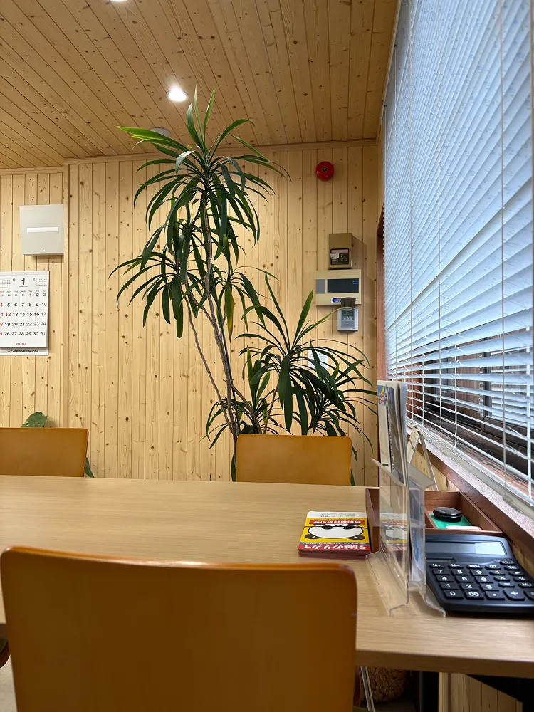 木目調の受付・デスク周り。観葉植物と壁面の設備（案内板、インターホンなど）が写るオフィス内観