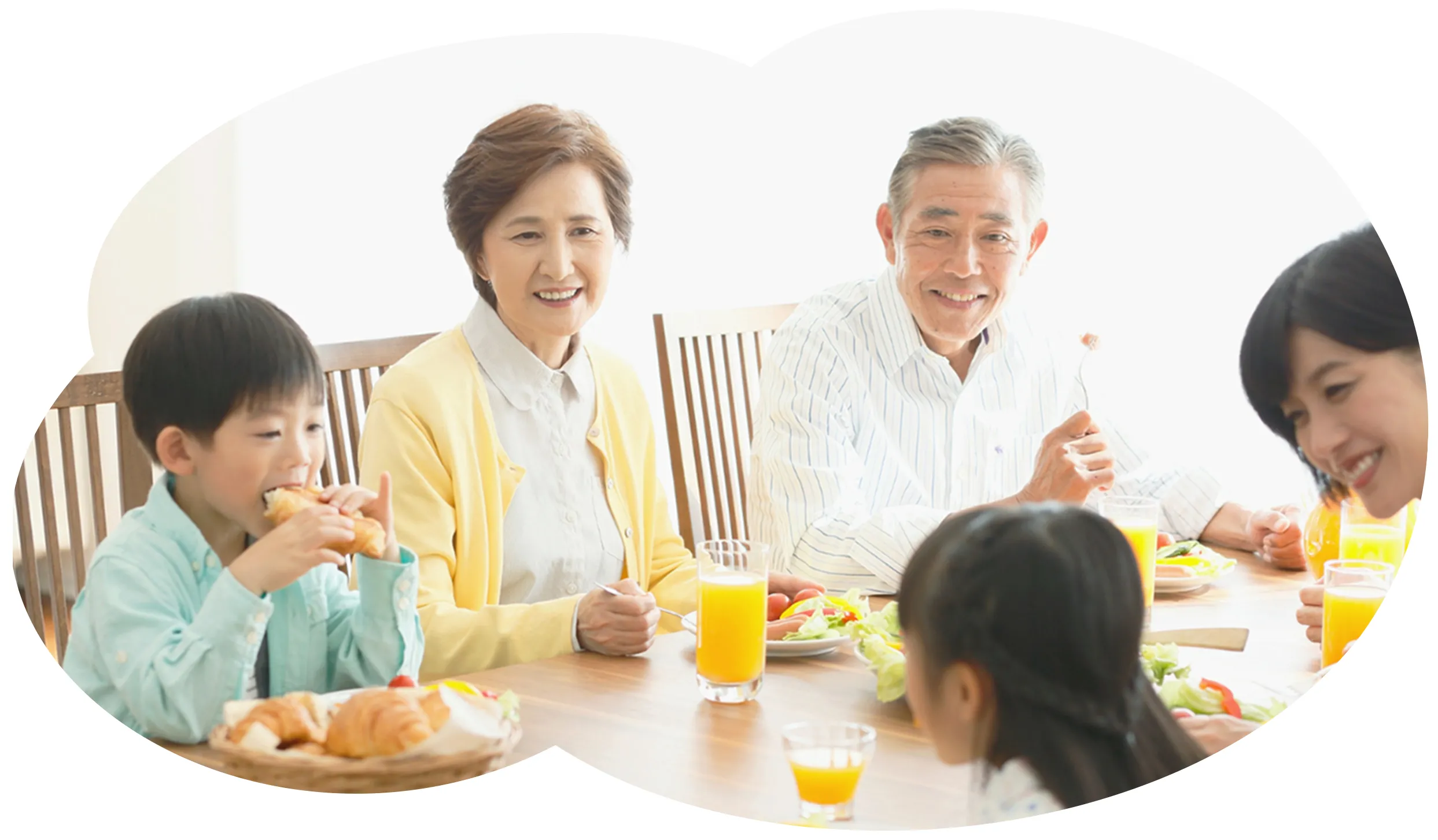 食卓で朝食を囲む家族（祖父母・両親・子ども2人、クロワッサンとオレンジジュース）