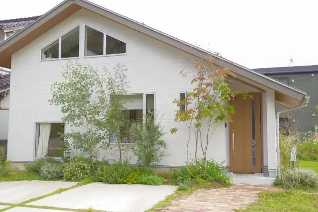 白いモダンな外観の一軒家。三角形の窓と木製玄関ドアが特徴。庭には植栽とアプローチ