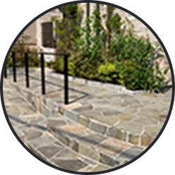 薄い色の石畳のパティオ、黒い金属手すり、緑の植栽（外構）